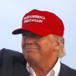 | 1776 | 1791 patriot shops close-up of a man wearing a red maga hat, smiling slightly, against a pale blue background.